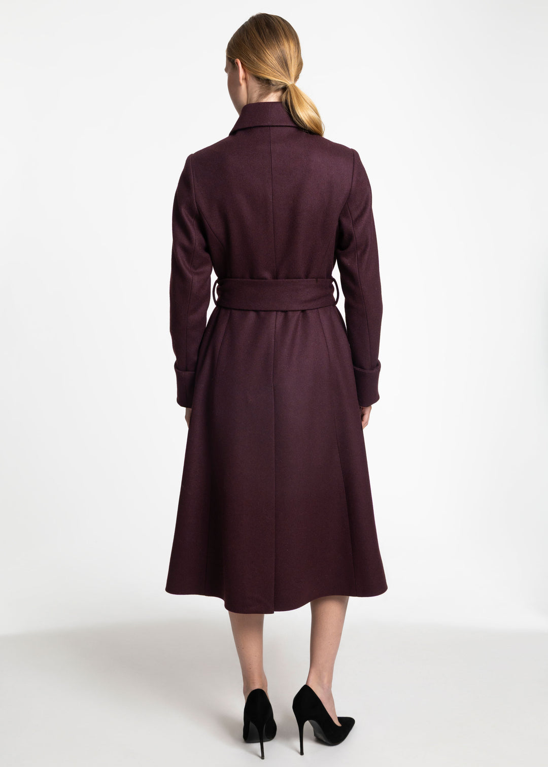 A woman viewed from the back wearing a knee-length dark plum coat with a matching waist tie and black high heels, against a plain white studio background.