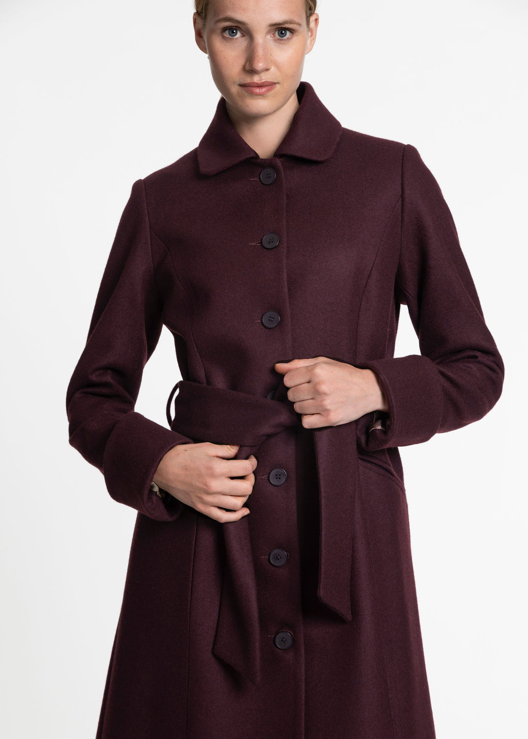A woman wearing a long, dark burgundy-plum wool coat with a collar, front buttons, and a matching waist tie, photographed against a plain light gray studio background.