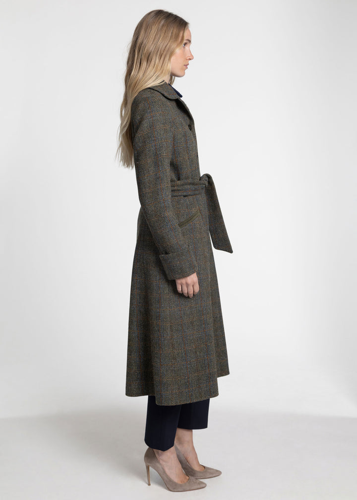 A woman modeling a long belted overcoat in side profile against a plain light gray studio background. The coat appears to be a dark olive-brown tweed with subtle plaid variation.