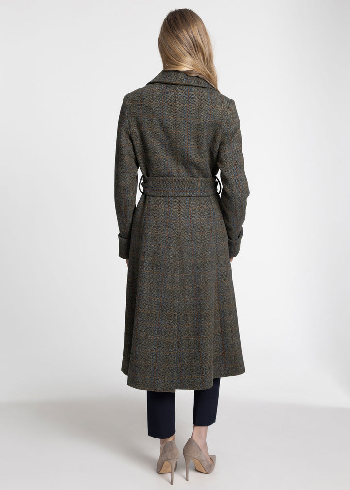 A woman viewed from the back wearing a long belted plaid coat in dark olive and brown tones, paired with dark pants and beige heels, against a plain light gray studio background.