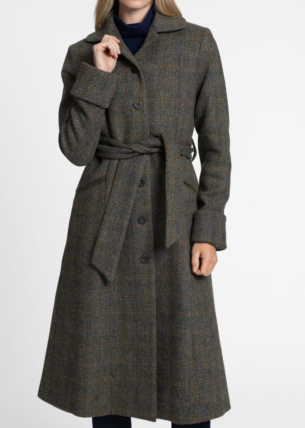 A woman wearing a long belted overcoat in a dark green plaid pattern, paired with a dark navy turtleneck, photographed against a plain white studio background.