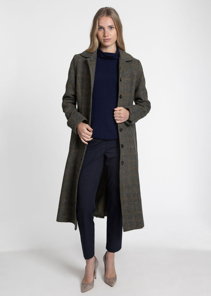 A woman modeling a long dark olive green and brown plaid coat over a navy blue turtleneck sweater, navy pants, and beige pointed-toe heels, photographed against a light gray studio background.