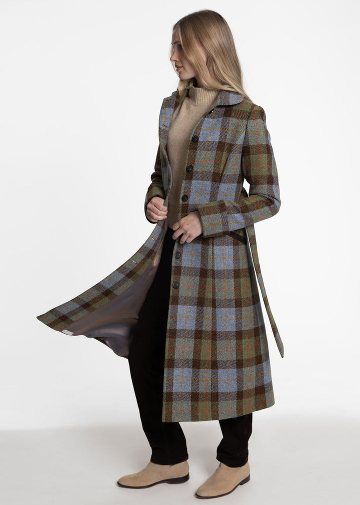 A woman modeling a long plaid coat in a studio setting. The coat features a green, brown, and blue tartan pattern with a beige turtleneck underneath and black pants.