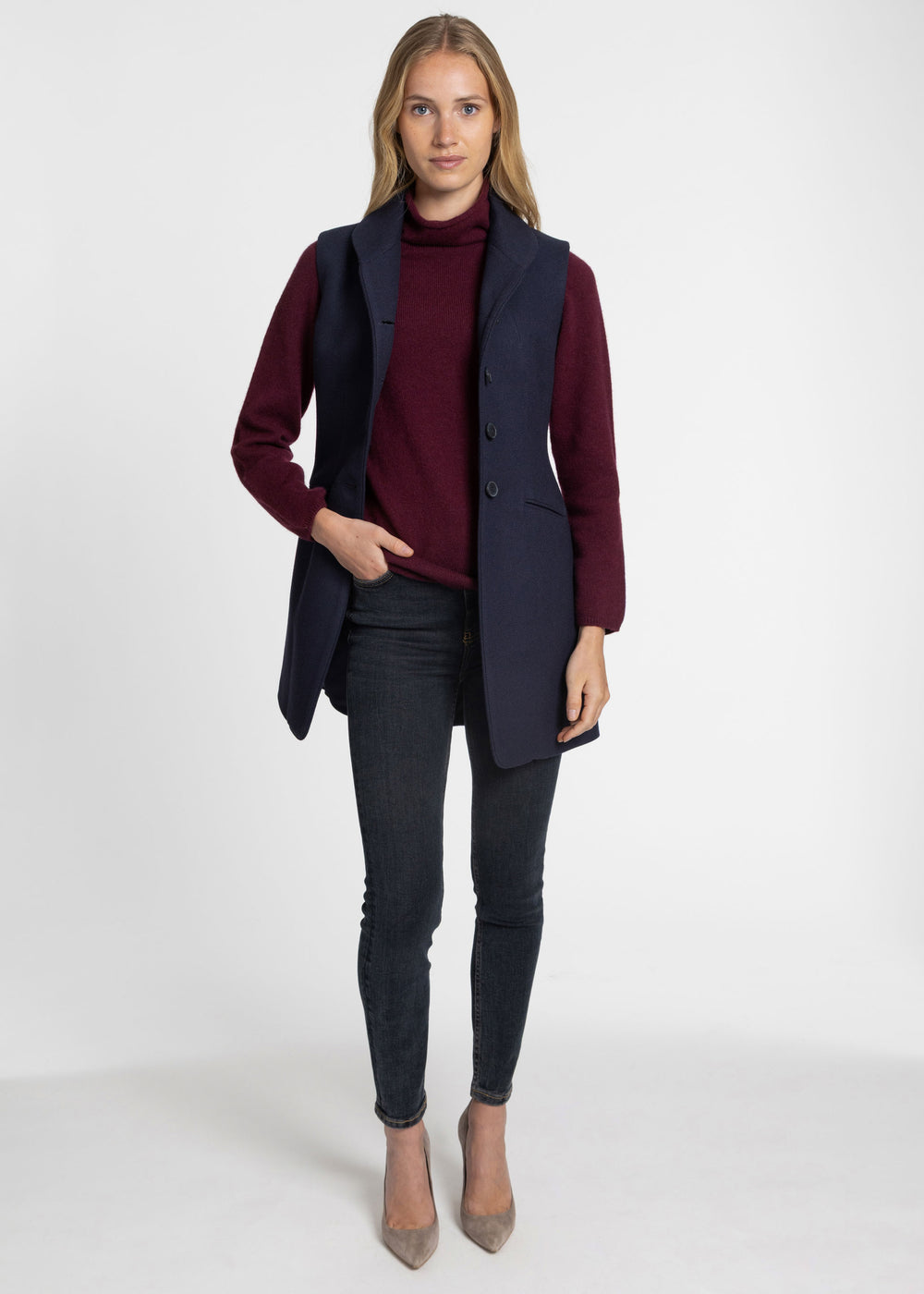 A woman modeling a dark navy sleeveless waistcoat over a burgundy turtleneck sweater, paired with dark gray skinny jeans and beige pointed-toe heels, against a plain white studio background.