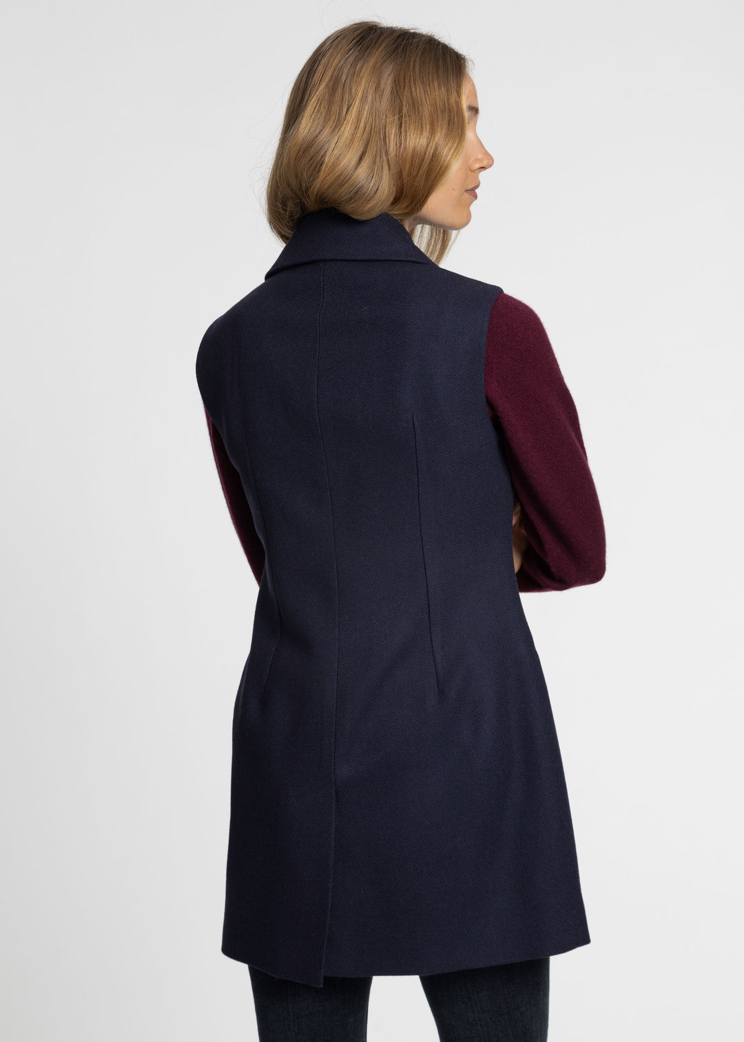 A woman shown from the back wearing a sleeveless dark navy waistcoat over a burgundy long-sleeve top, photographed against a plain light gray studio background.
