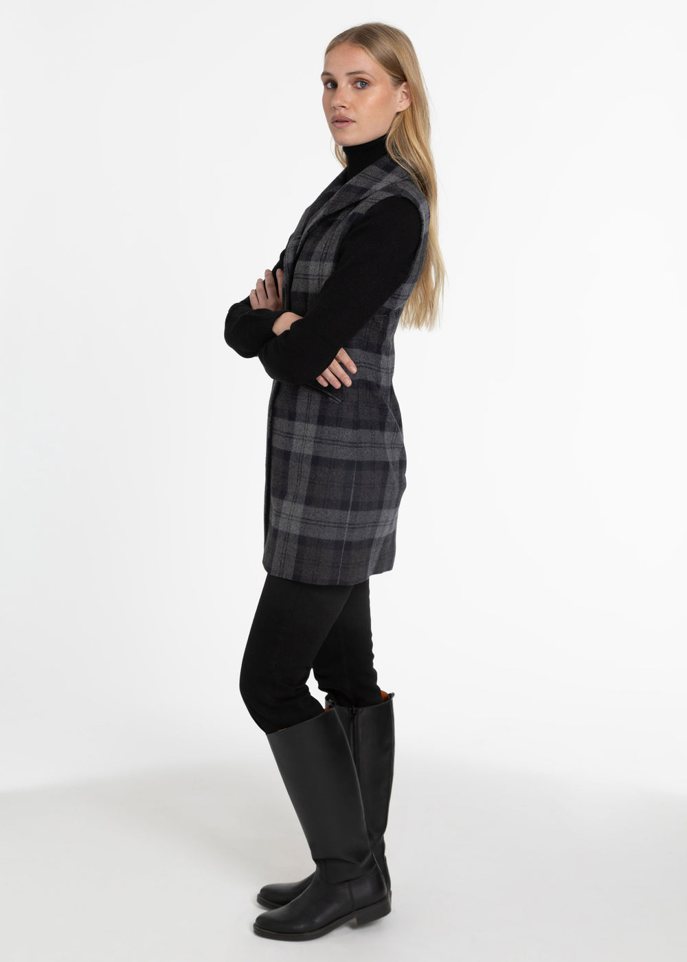A woman modeling a black and gray plaid sleeveless waistcoat over a black turtleneck, paired with black pants and tall black boots, standing against a plain white studio background.