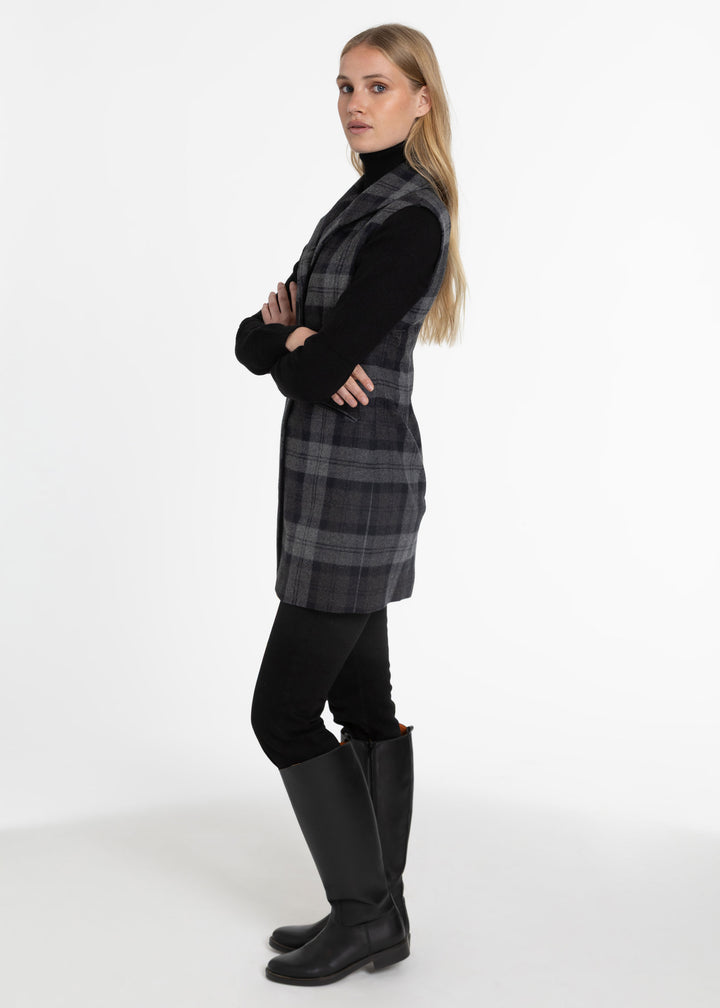 A woman modeling a black and gray plaid sleeveless waistcoat over a black turtleneck, paired with black pants and tall black boots, standing against a plain white studio background.