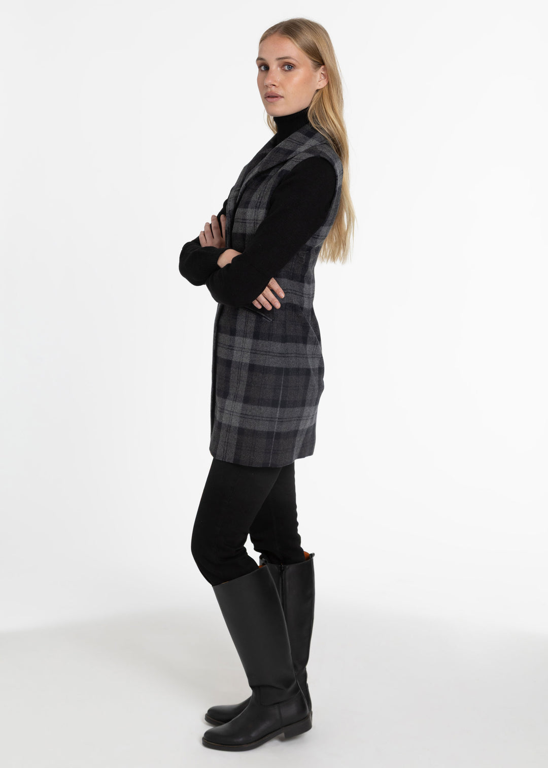 A woman modeling a black and gray plaid sleeveless waistcoat over a black turtleneck, paired with black pants and tall black boots, standing against a plain white studio background.