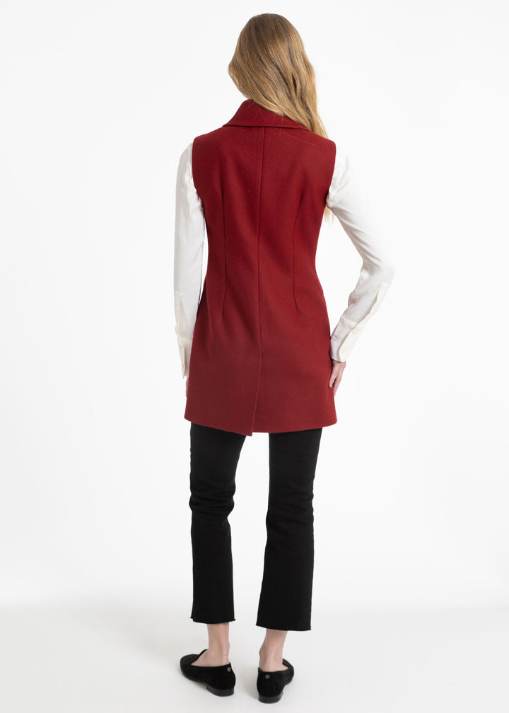 A woman shown from the back wearing a sleeveless red waistcoat over a white long-sleeve shirt, black cropped pants, and black flats, against a plain white studio background.