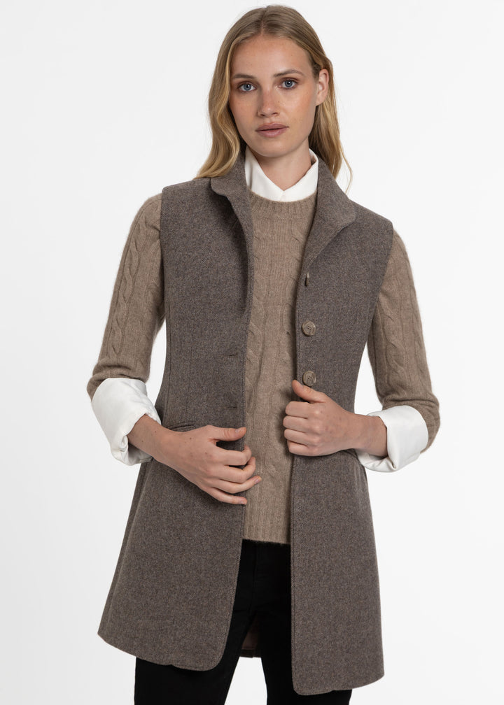A woman modeling a taupe-brown sleeveless wool waistcoat over a beige cable-knit sweater and white collared shirt, against a plain white studio background.
