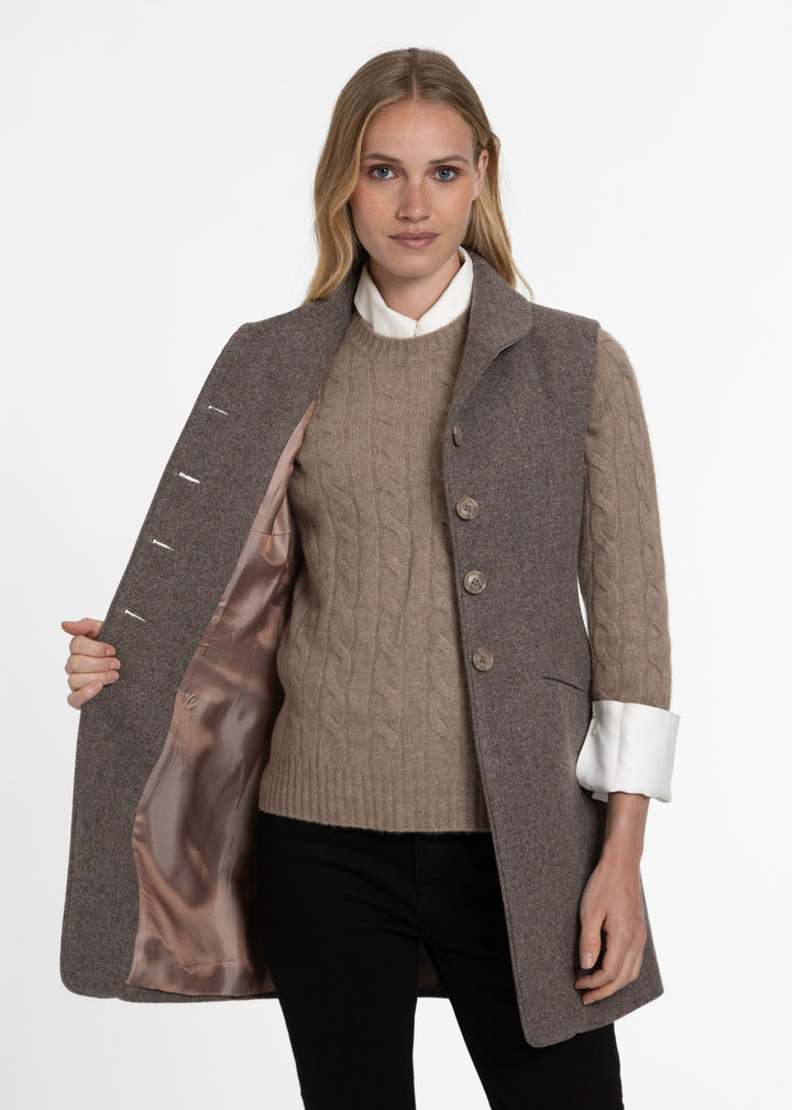 A woman modeling a brown wool-blend waistcoat with a satin lining, worn over a beige cable-knit sweater and a white collared shirt, against a plain white studio background.