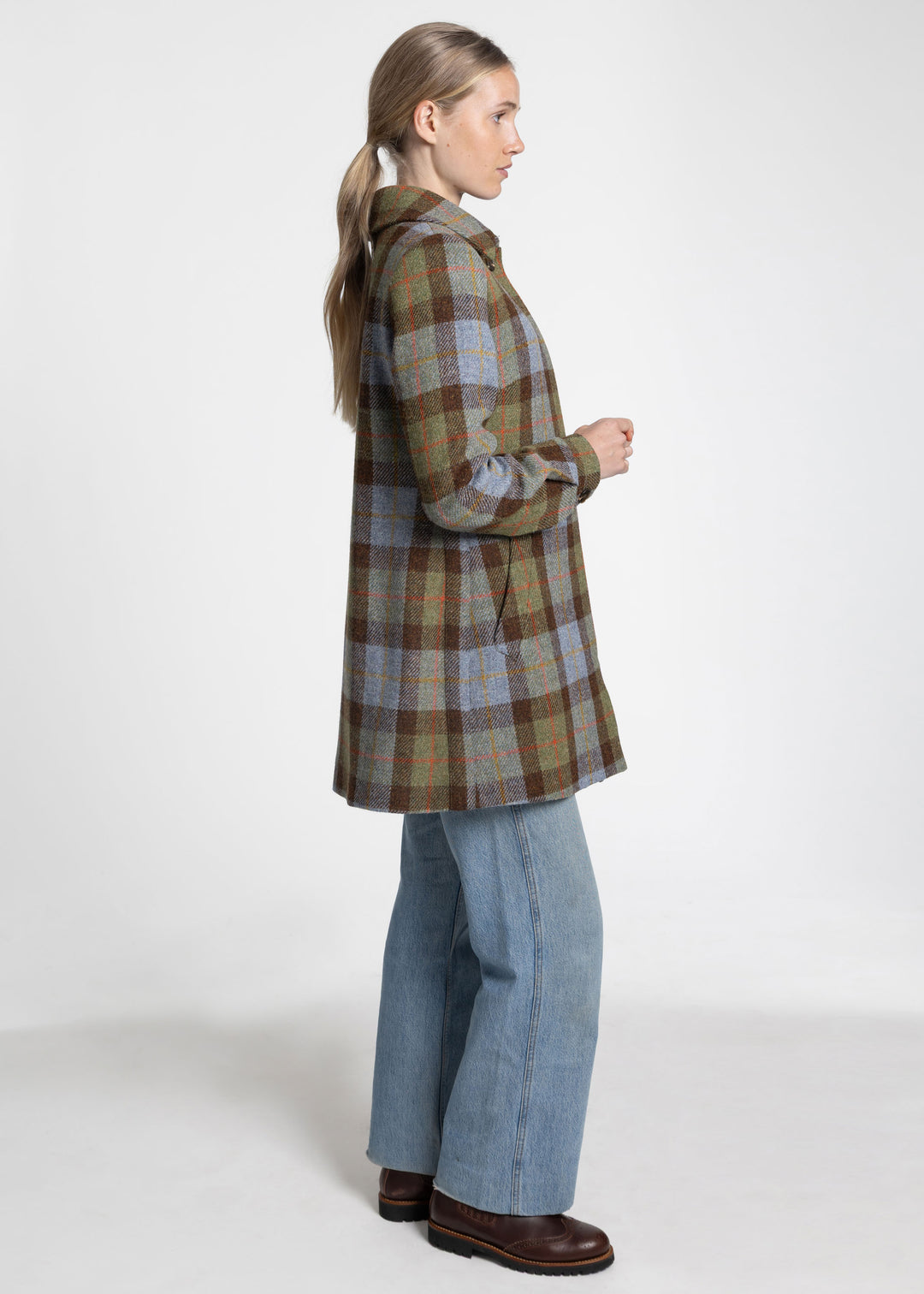 A woman shown in profile wearing a brown, green, and blue plaid coat over a light blue wide-leg denim jacket and brown boots, photographed against a plain white studio background.