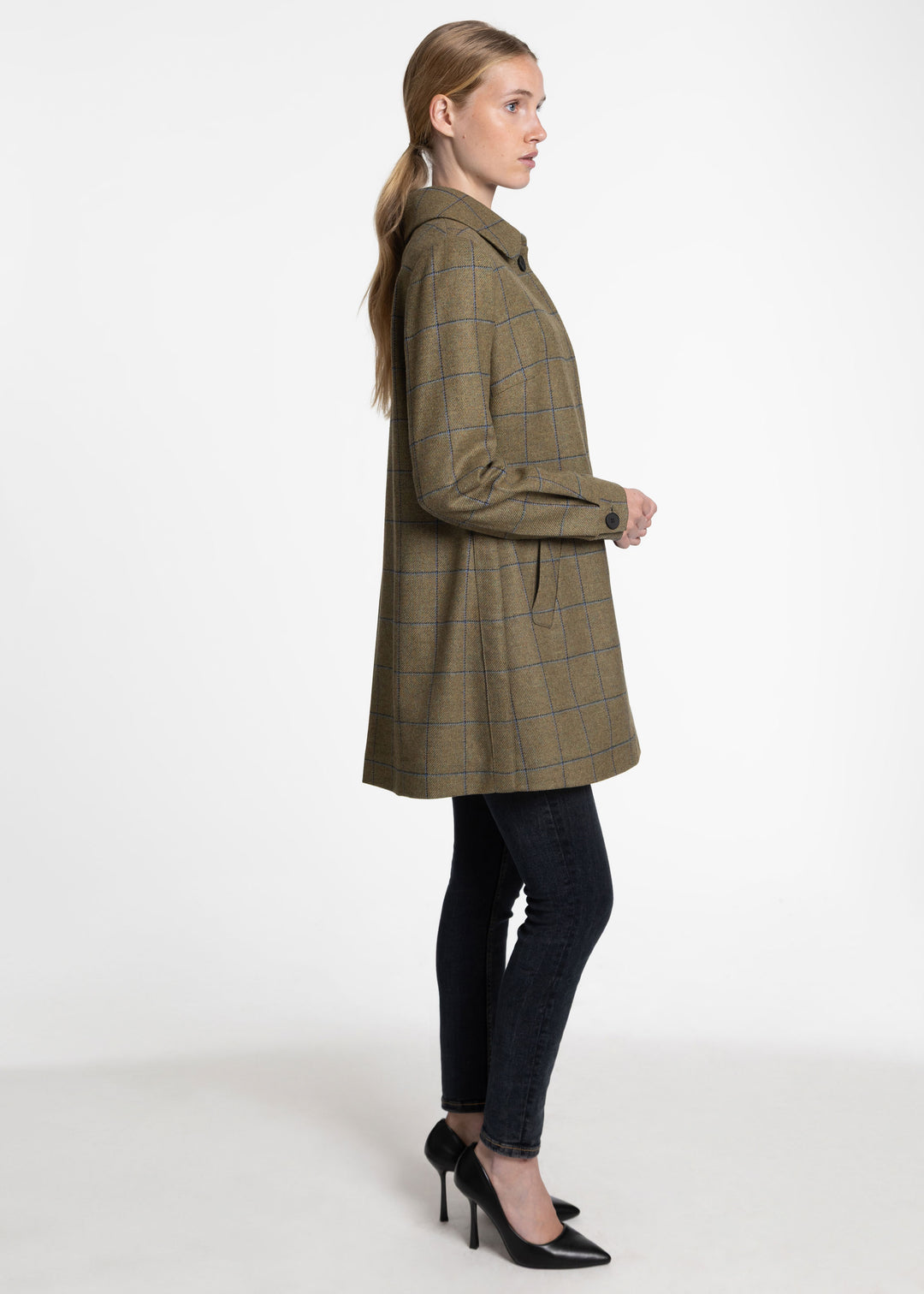 A woman modeling a mid-length plaid coat in side profile against a plain white studio background. The coat is olive green with a subtle blue windowpane plaid pattern, paired with dark skinny jeans and black pointed-toe heels.