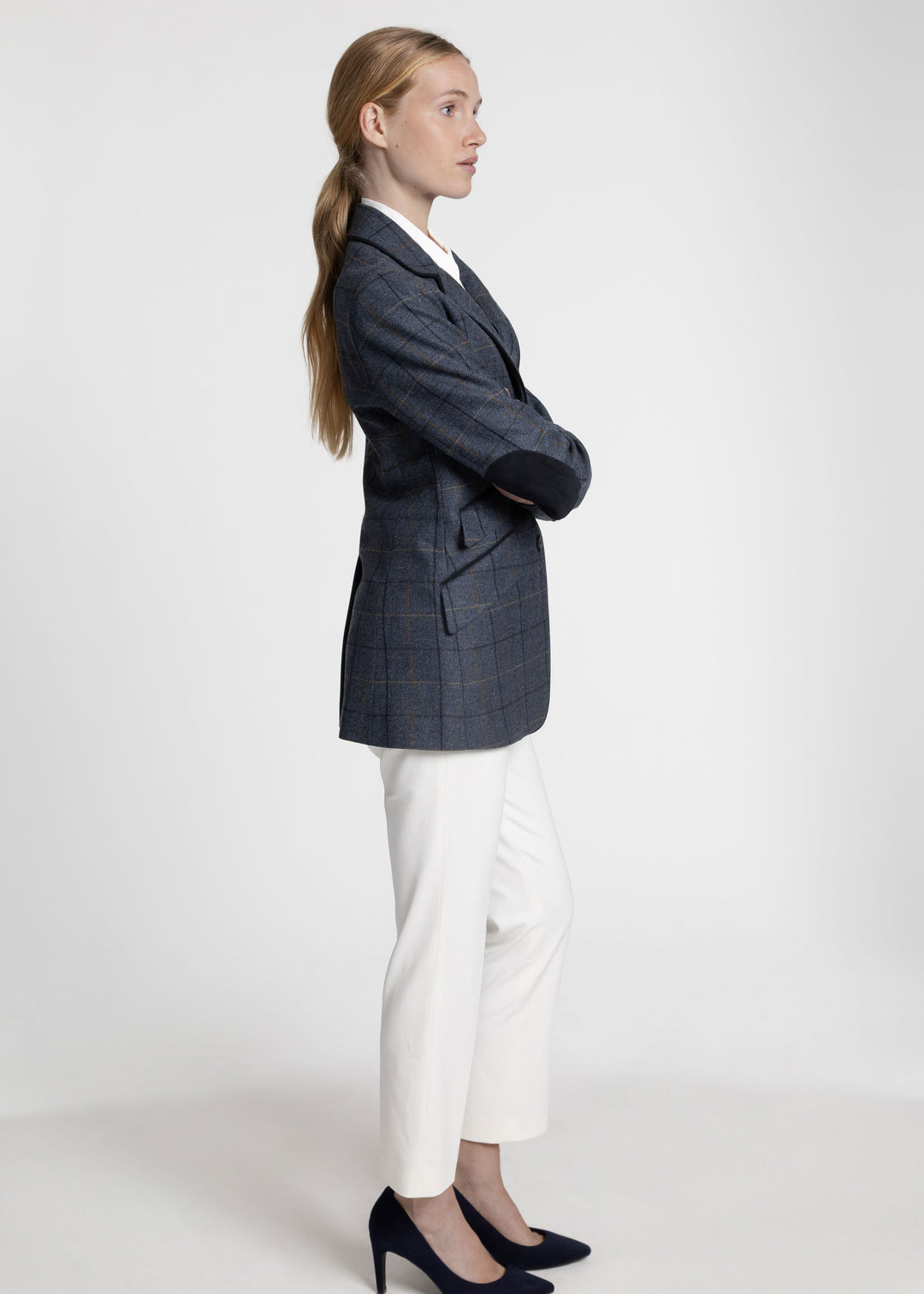 A woman in profile wearing a dark navy blue plaid blazer with navy elbow patches, white trousers, and navy heels, standing against a light gray studio background.