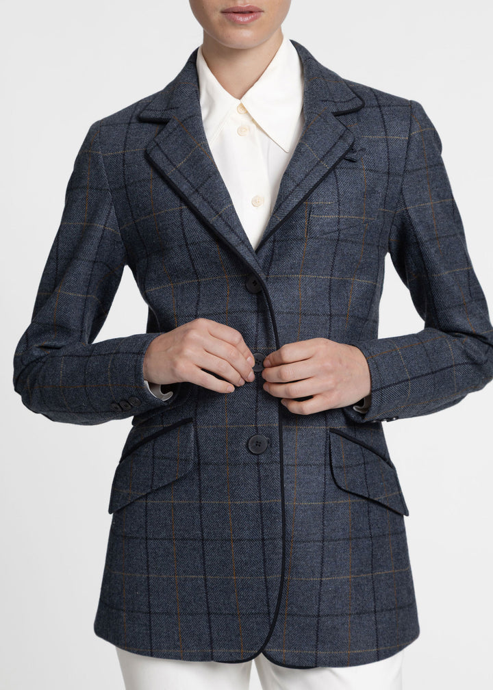 A person wearing a tailored jacket in dark blue with a subtle tan windowpane plaid pattern, paired with a white collared shirt and white trousers, against a light gray studio background.