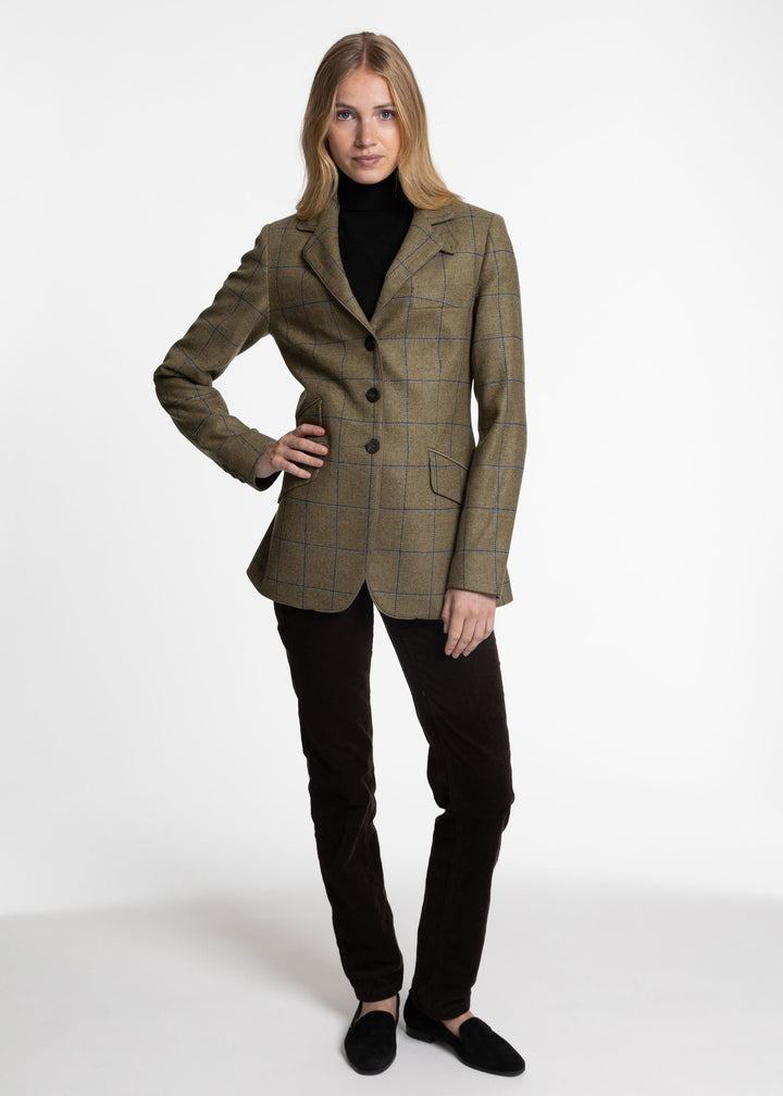A woman modeling a tailored jacket in olive green with a subtle windowpane check pattern, worn over a black turtleneck, black trousers, and black slip-on shoes against a plain white studio background.