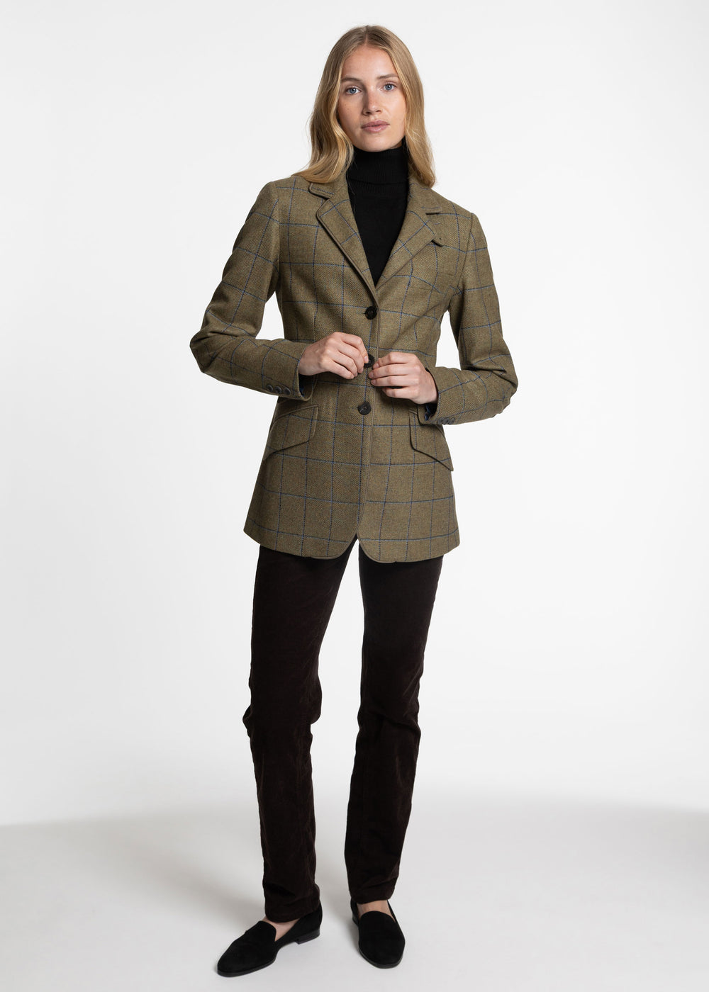 A woman modeling a tailored blazer in a green windowpane check pattern, worn over a black turtleneck and dark trousers, against a plain white studio background.
