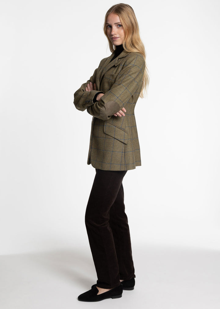 A woman modeling a tailored plaid jacket in olive green and brown tones, paired with dark brown trousers and black slip-on shoes, against a plain white studio background.