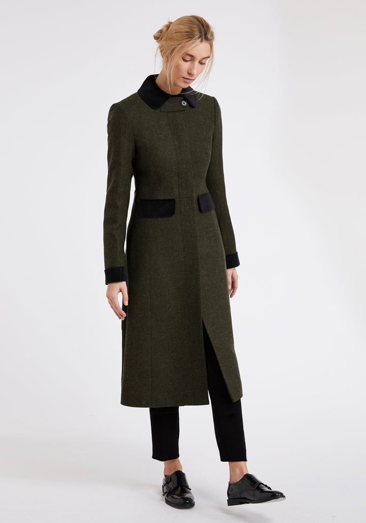 A woman modeling a long olive green wool coat with black collar, cuffs, and pocket details, paired with black pants and shoes, against a plain white studio background.