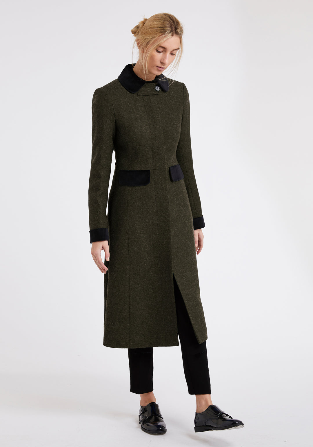 A woman modeling a long olive green wool coat with black collar, cuffs, and pocket details, paired with black pants and shoes, against a plain white studio background.
