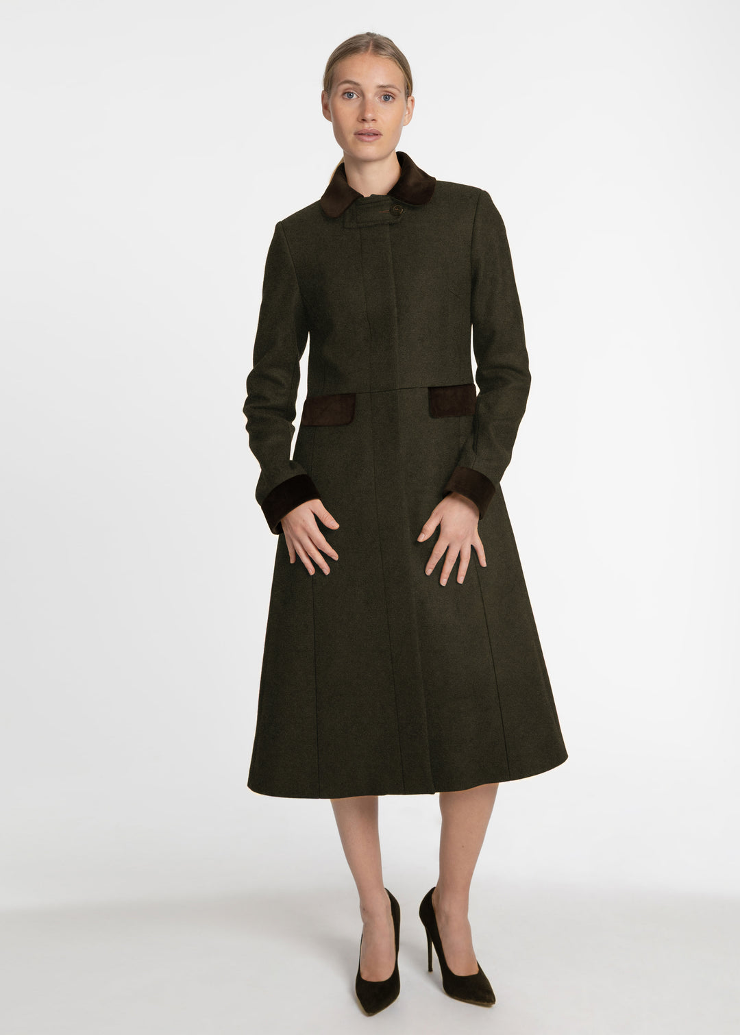 A woman modeling a dark olive green knee-length coat with contrasting dark brown collar, cuffs, and pocket details, paired with black heels, against a plain light gray studio background.