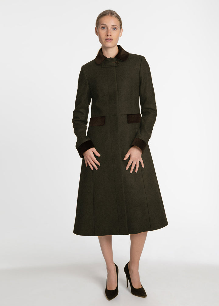 A woman modeling a dark olive green knee-length coat with contrasting dark brown collar, cuffs, and pocket details, paired with black heels, against a plain light gray studio background.