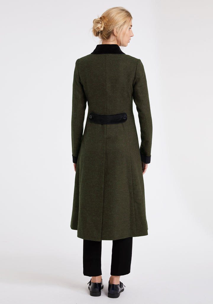 Back view of a woman wearing a dark olive green wool coat with black velvet details on the back belt, collar and cuffs. Worn with black trousers and dress shoes.