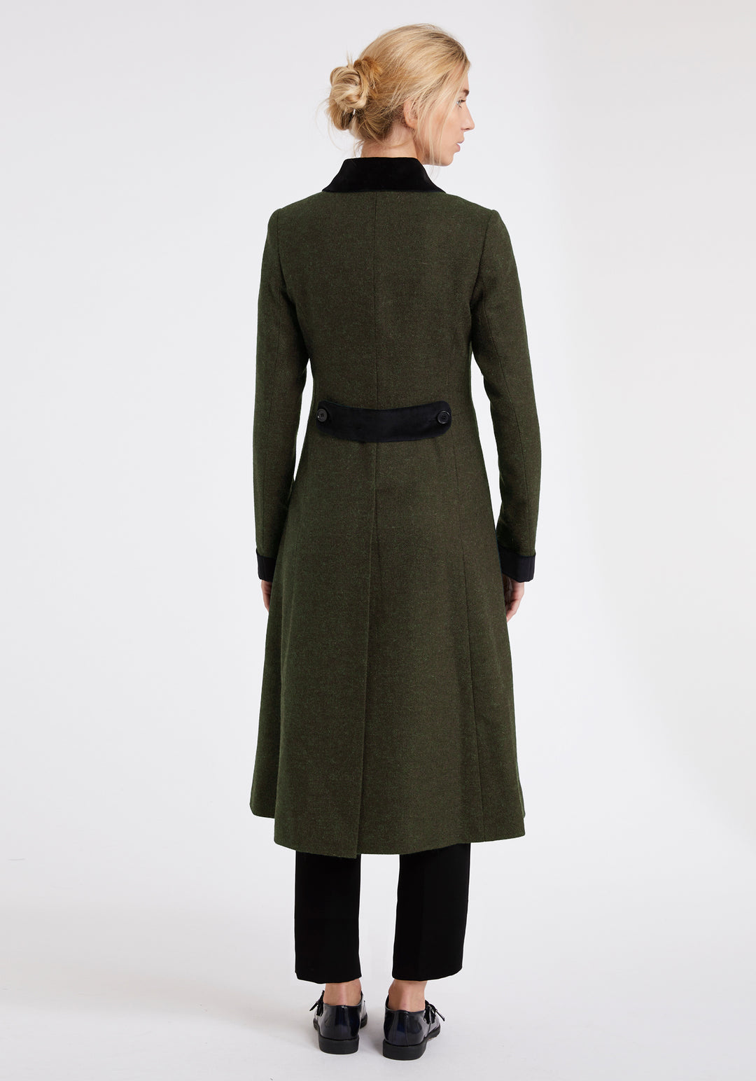 Back view of a woman wearing a dark olive green wool coat with black velvet details on the back belt, collar and cuffs. Worn with black trousers and dress shoes.