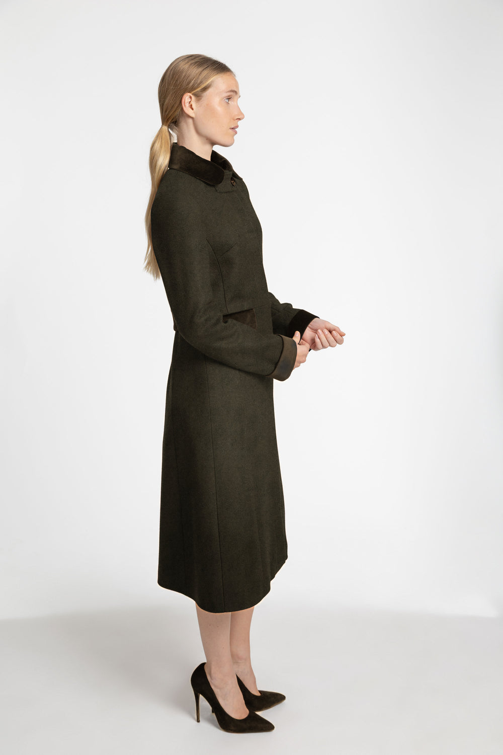 A woman in profile wearing a dark olive green knee-length coat with a high dark collar and black pointed-toe heels, standing against a plain light gray studio background.