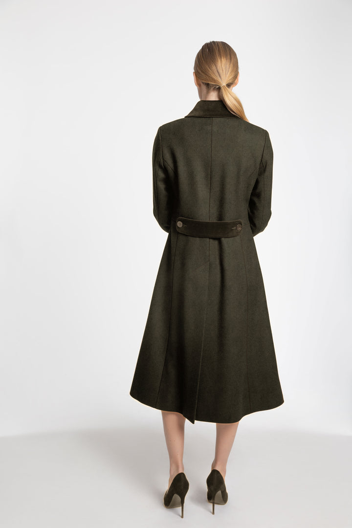 A woman viewed from the back wearing a dark olive green knee-length coat with a matching velvet belt, paired with dark pointed-toe heels, against a plain light gray studio background.