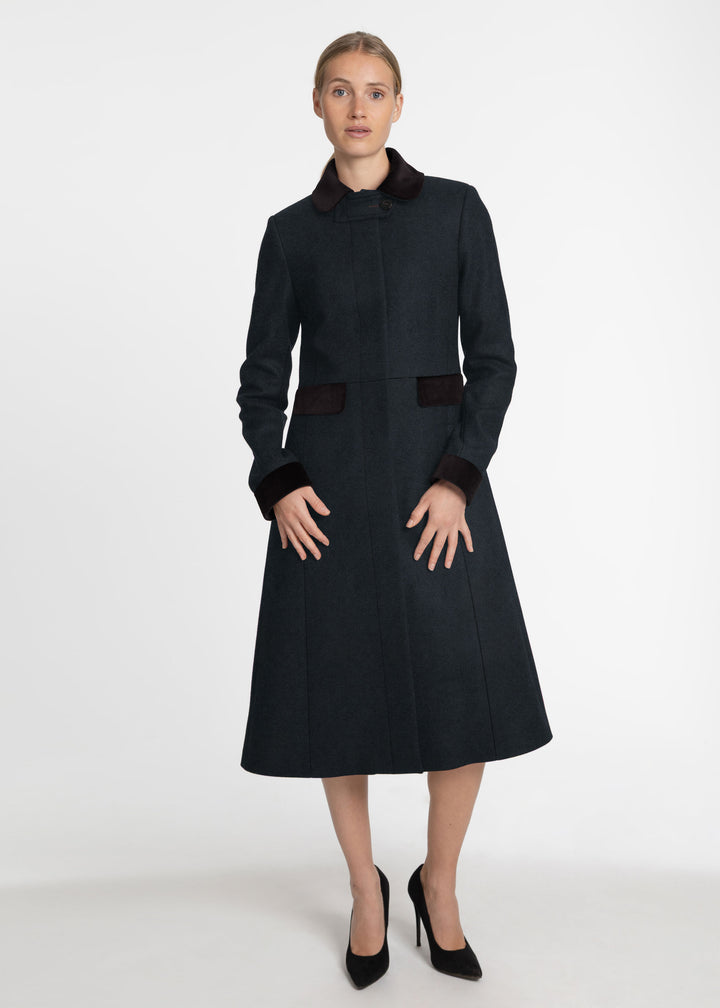 A woman modeling a dark green wool coat with contrasting dark black collar, cuffs, and waist detail, paired with black high heels, against a plain white studio background.