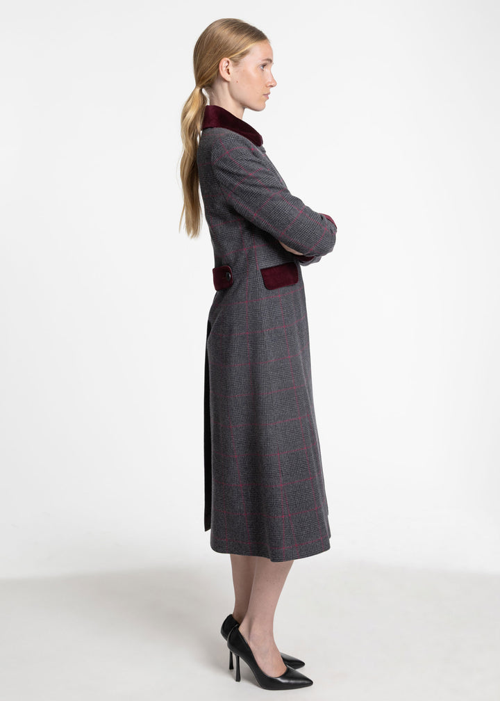 A woman modeling a long gray plaid coat with dark red velvet trim at the collar and waist, shown in profile against a plain white studio background.