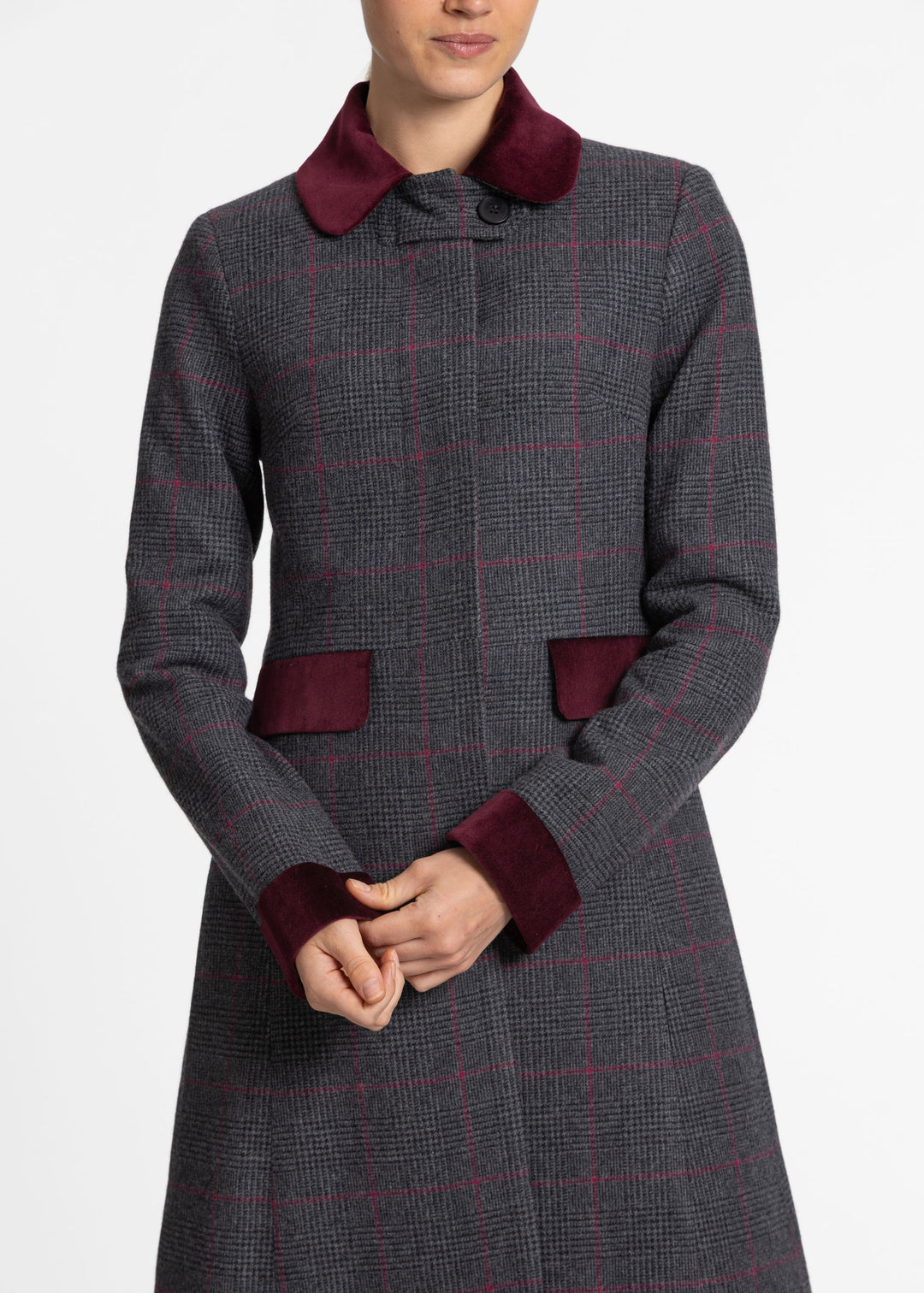 A woman wearing a long gray windowpane coat with dark red velvet collar, cuffs, and pocket flaps, shown against a plain light gray studio background.