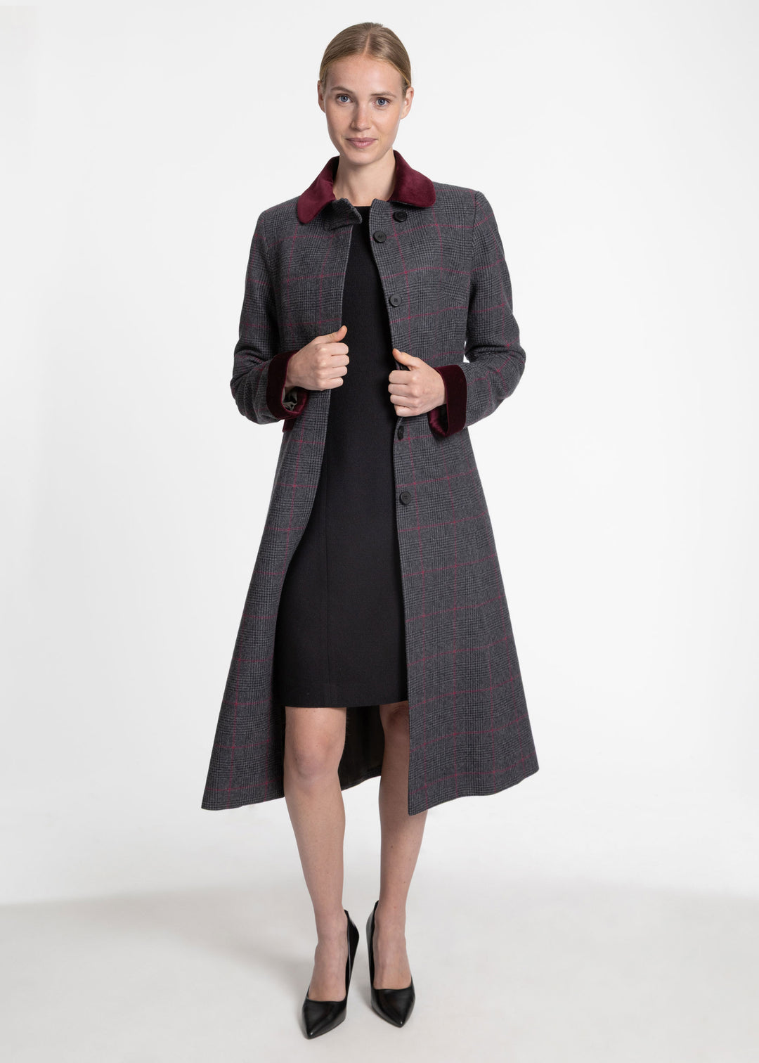 A woman modeling a long charcoal gray plaid coat with a dark burgundy velvet collar and cuffs, worn over a black dress and black pointed-toe heels, against a plain white studio background.