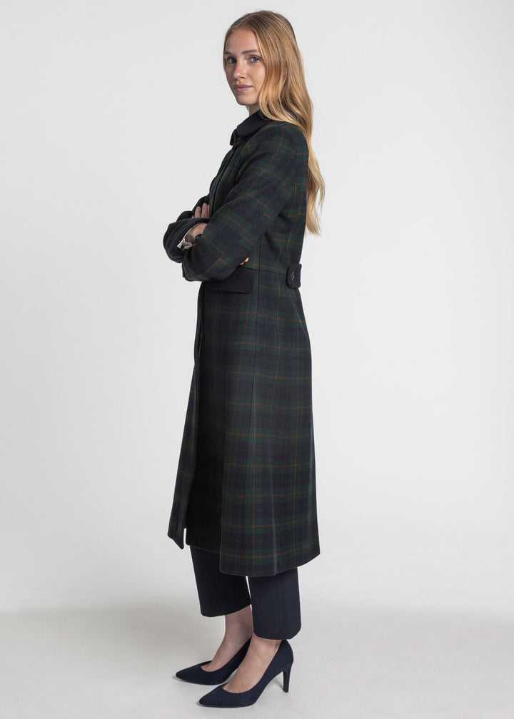 A woman modeling a long dark green and navy plaid coat, paired with dark trousers and navy heels, standing against a plain light gray studio background.