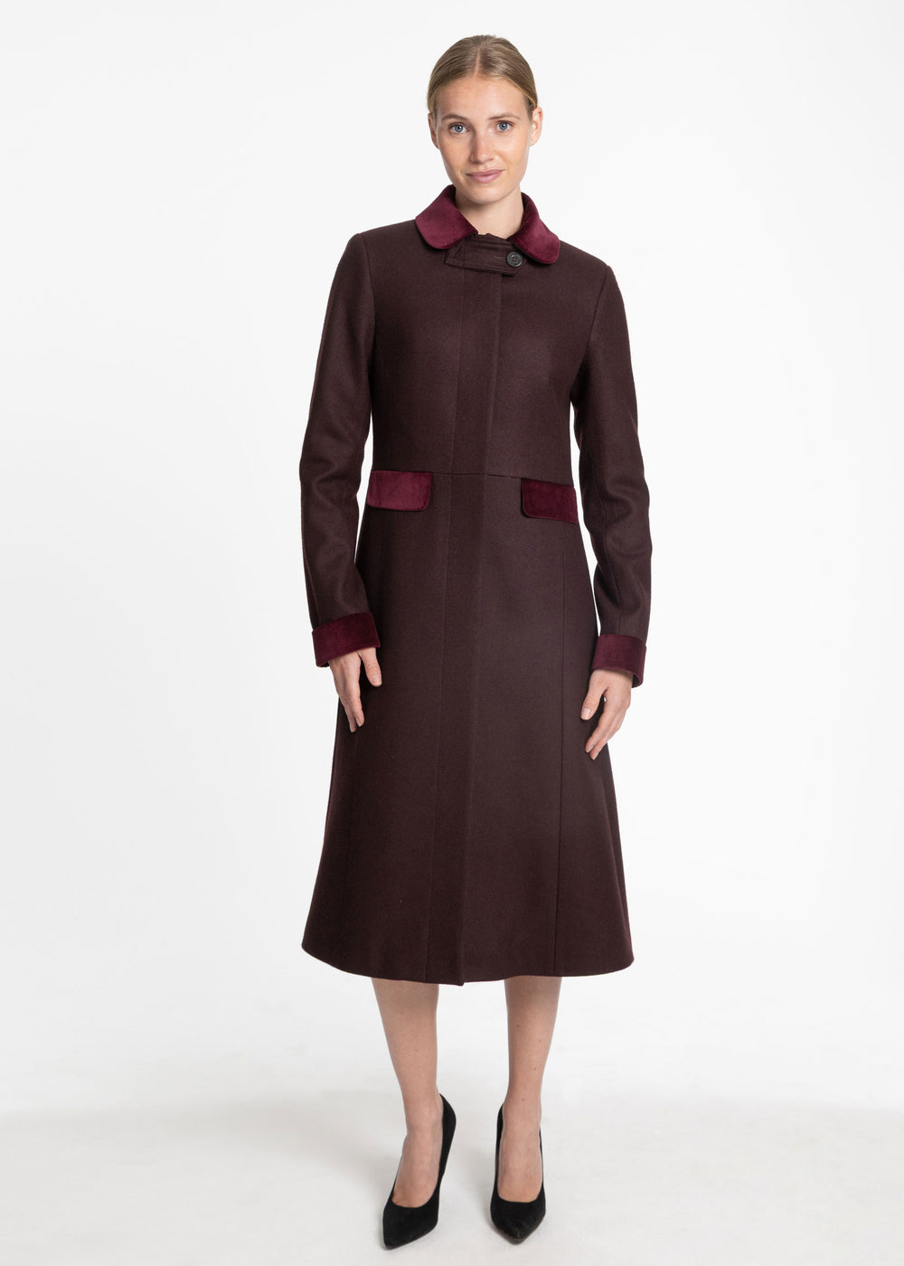A woman modeling a knee-length dark mulberry coat with contrasting burgundy collar, cuffs, and waist detail, paired with black heels, against a plain white studio background.