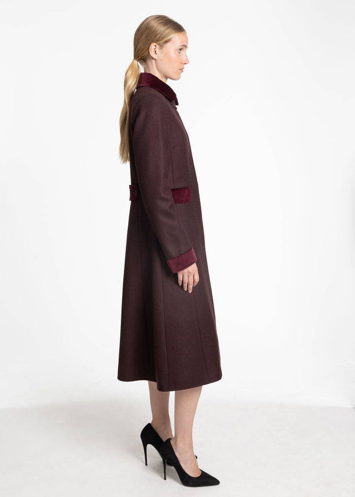 A woman modeling a long dark mulberry wool coat with contrasting burgundy velvet collar, cuffs, and pocket details, paired with black high heels against a plain white studio background.
