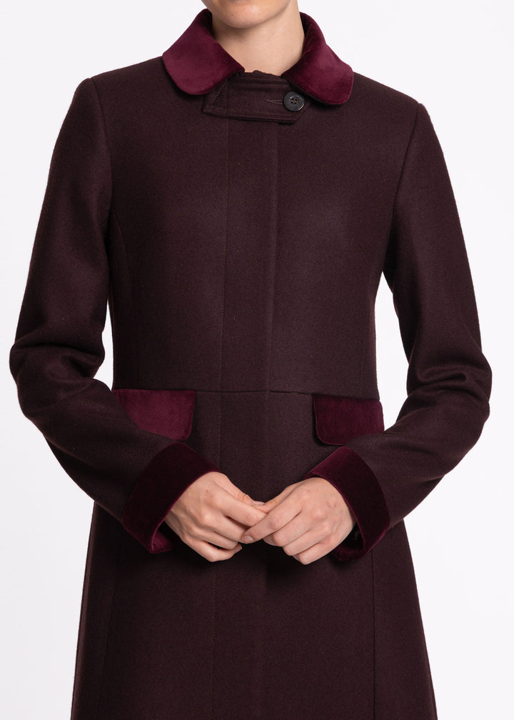 A close-up of a model wearing a dark mulberry wool coat with contrasting deep burgundy velvet collar, cuffs, and pocket flaps.