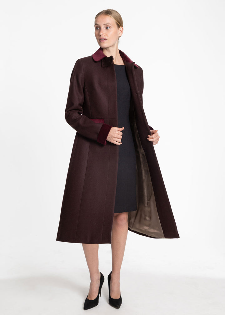 A woman models a long dark mulberry wool coat with contrasting burgundy velvet lapels and cuffs, worn open over a black dress and black heels, against a plain white studio background.