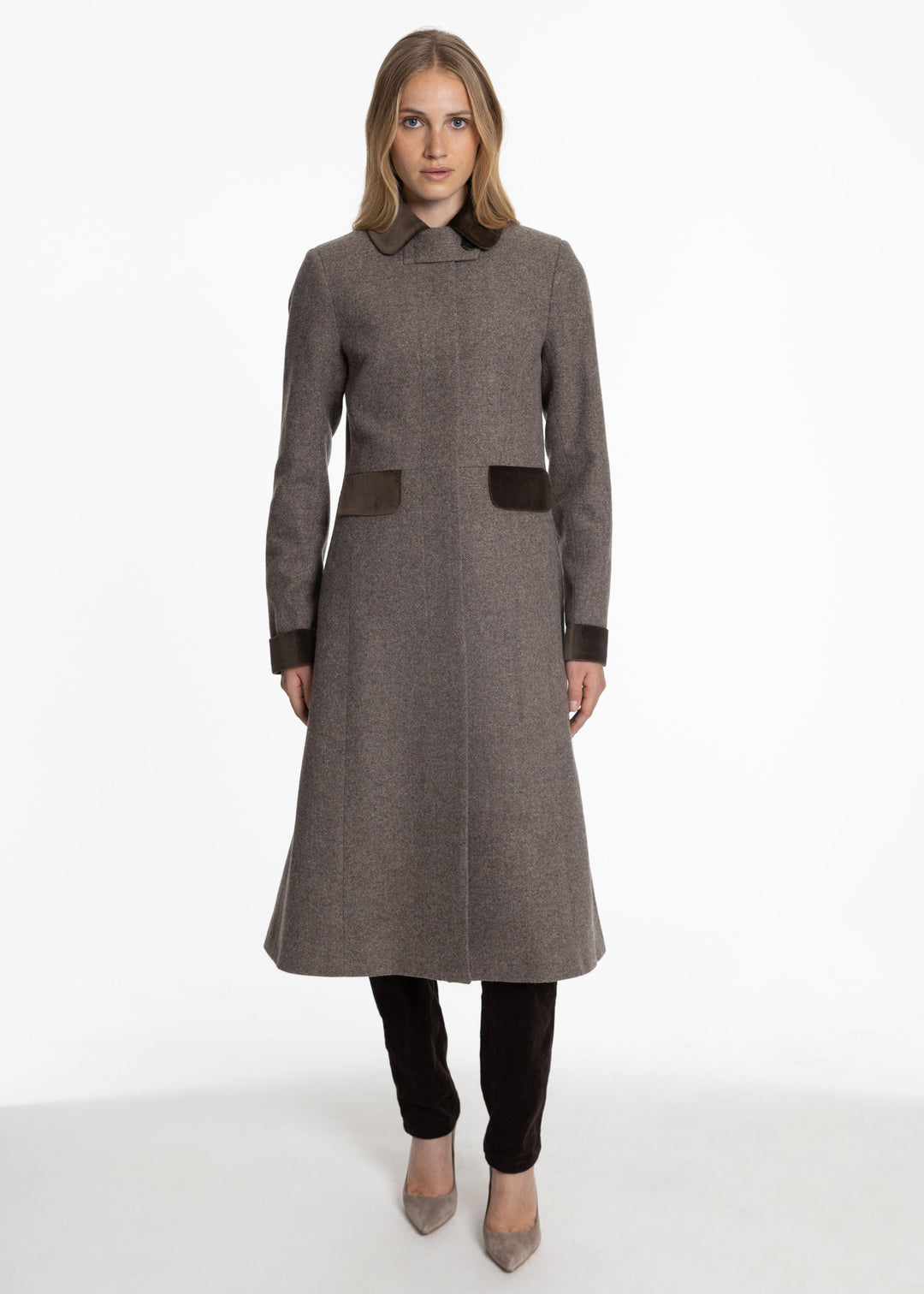 A woman modeling a mid-length brown wool coat with dark brown contrast trim on the collar, cuffs, and front pockets, paired with black pants and beige heels against a white studio background.