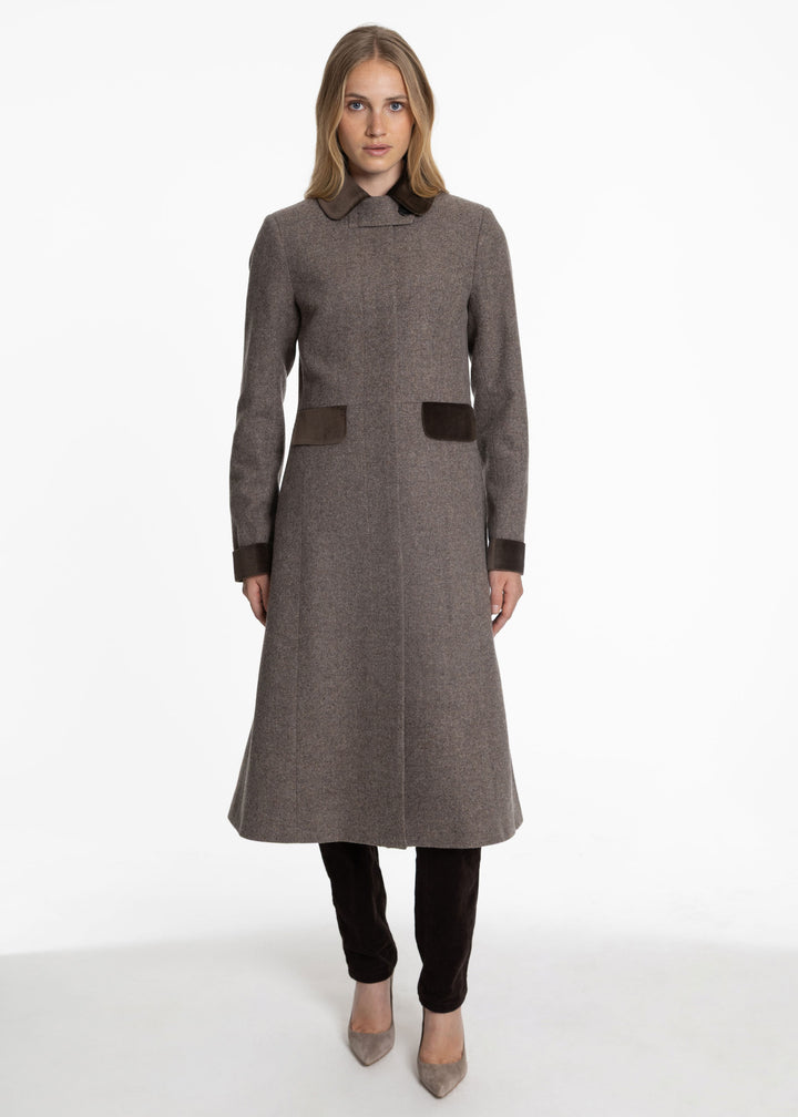 A woman modeling a mid-length brown wool coat with dark brown contrast trim on the collar, cuffs, and front pockets, paired with black pants and beige heels against a white studio background.
