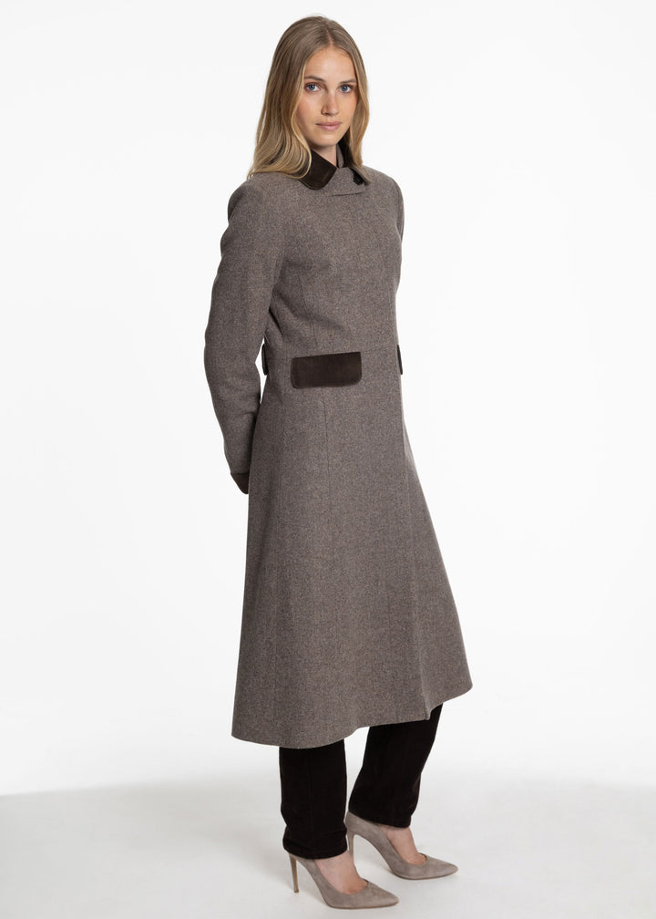 A woman modeling a long purple-brown wool-blend coat with dark brown trim details, including a collar, waist belt, and front pocket, paired with black pants and beige heels against a white studio background.