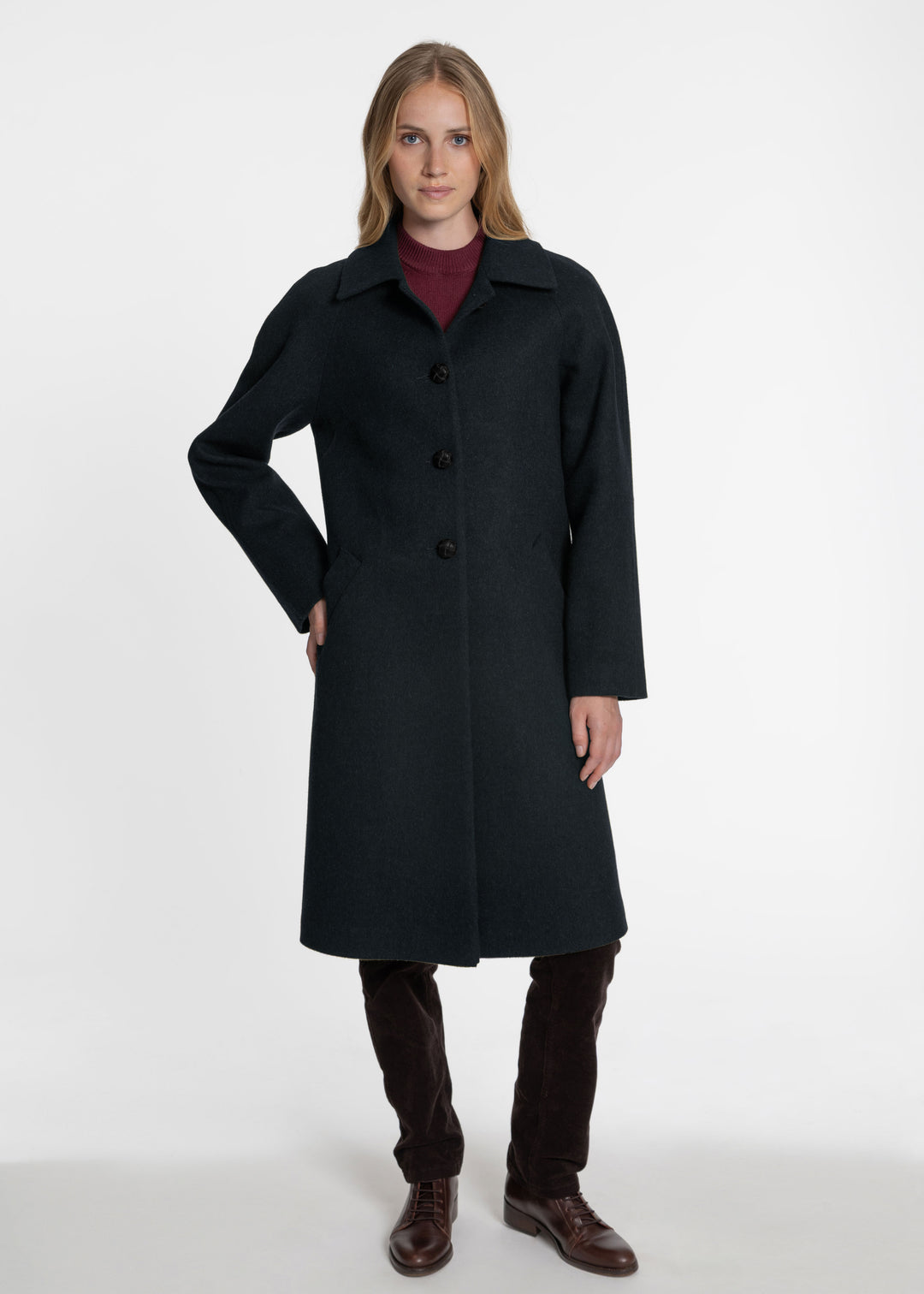 A woman modeling a long dark green wool-blend coat over a burgundy turtleneck, dark brown trousers, and brown leather shoes, photographed against a seamless white studio background.