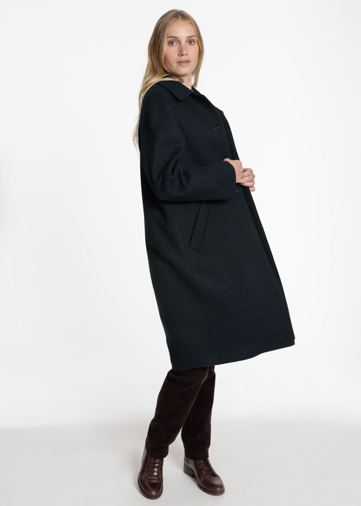 A woman modeling a long dark green coat, paired with dark brown trousers and brown leather shoes, against a plain white studio background.