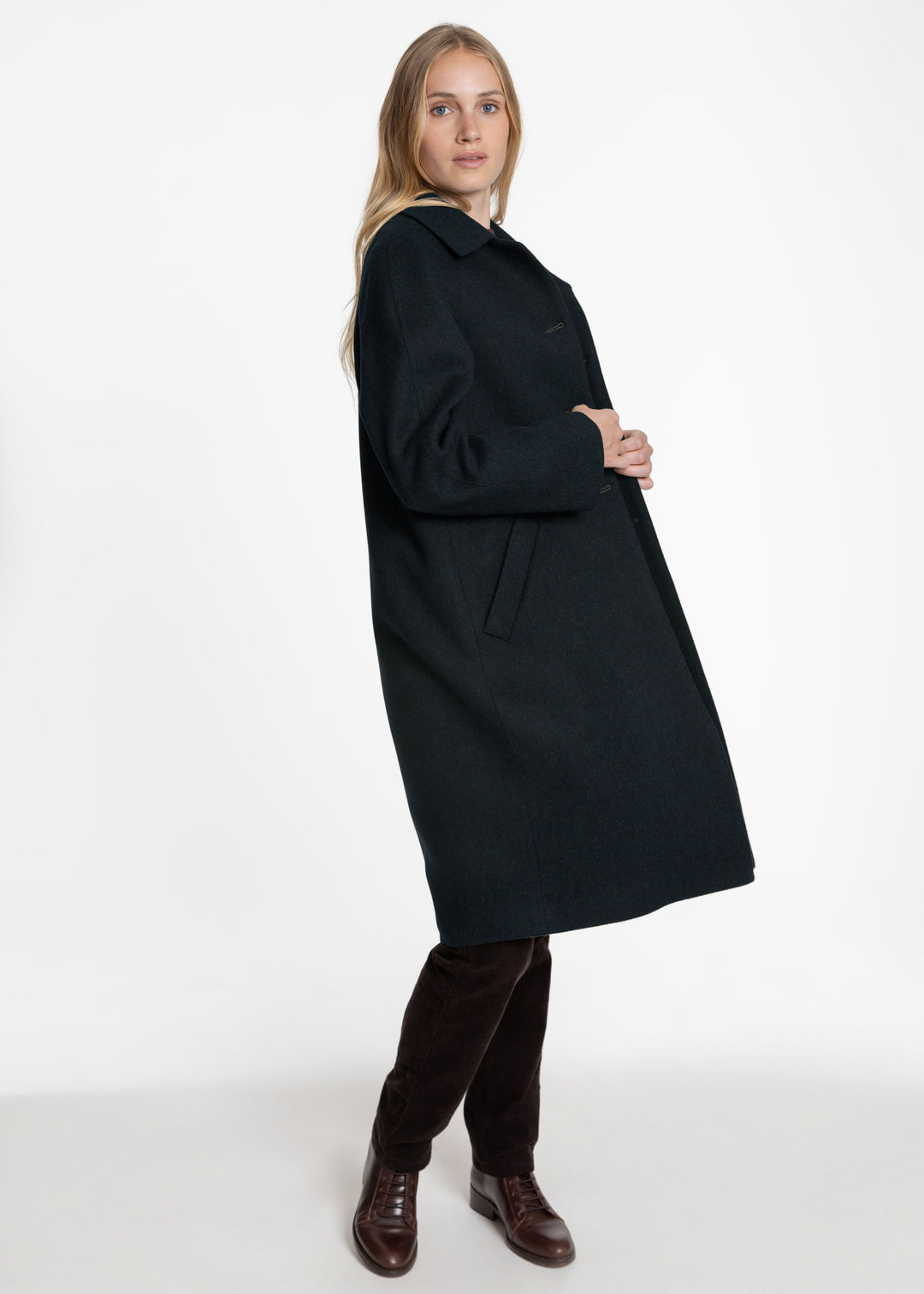A woman modeling a long dark green coat, paired with dark brown trousers and brown leather shoes, against a plain white studio background.