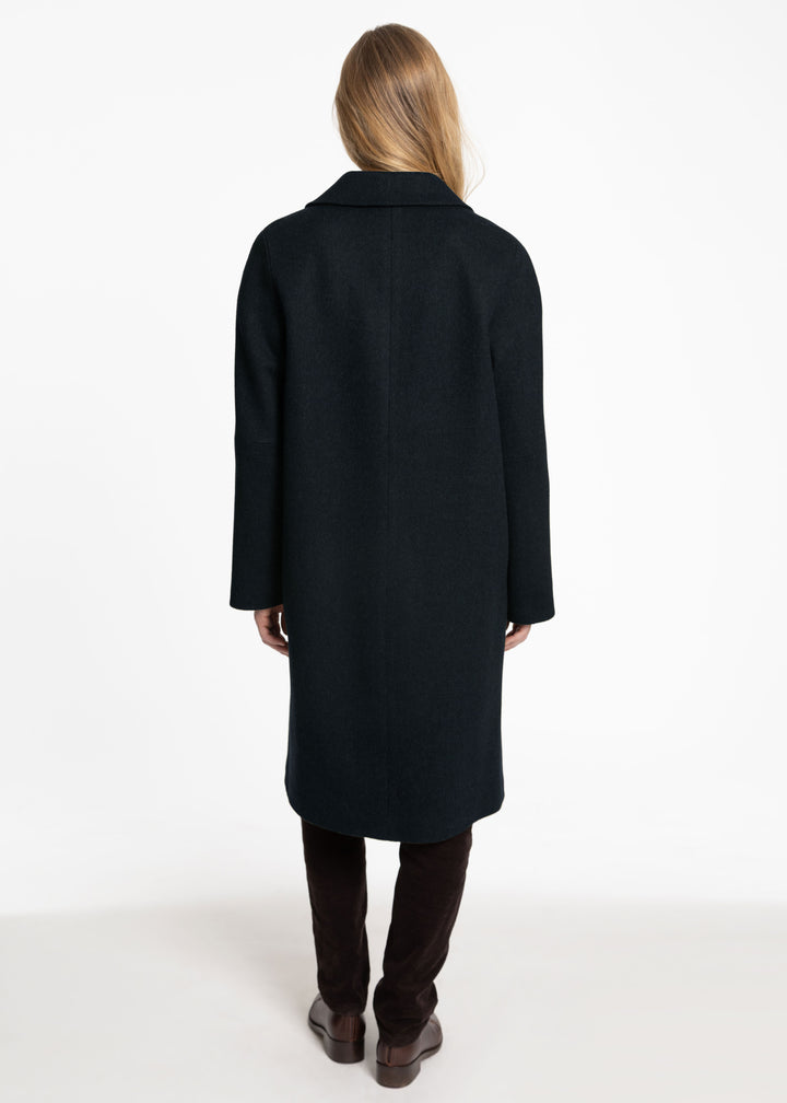 A woman viewed from the back wearing a long dark green coat, dark brown pants, and brown boots against a plain white studio background.