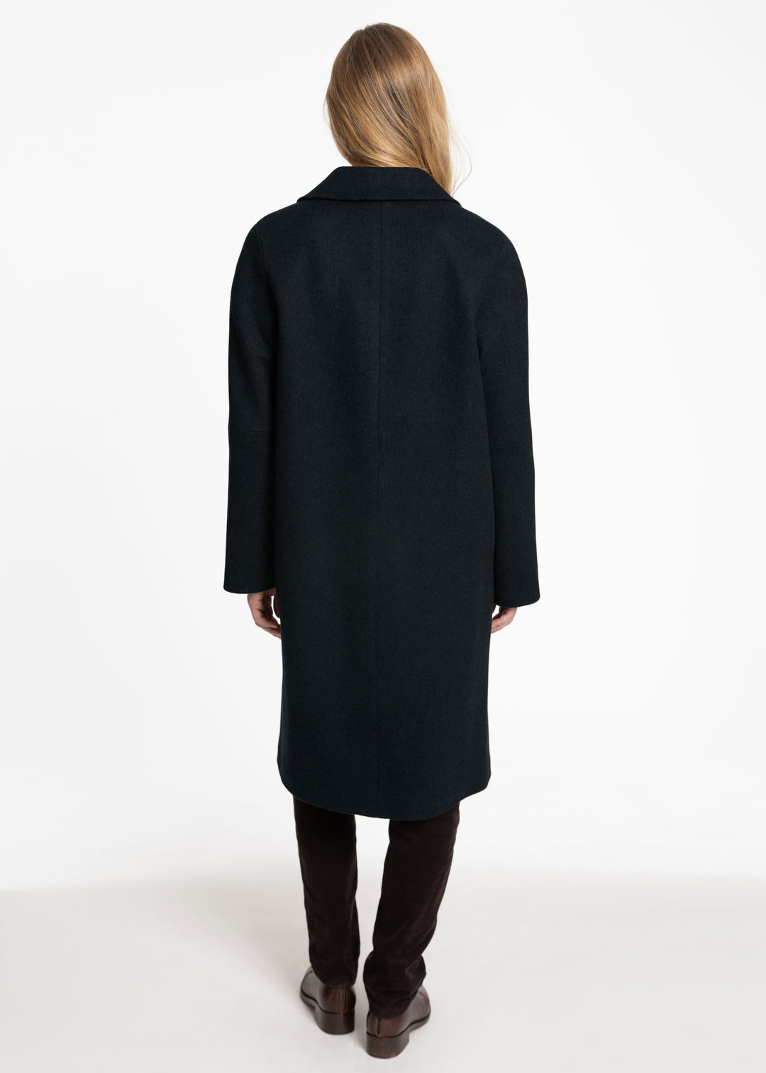 A woman viewed from the back wearing a long dark green coat, dark brown pants, and brown boots against a plain white studio background.