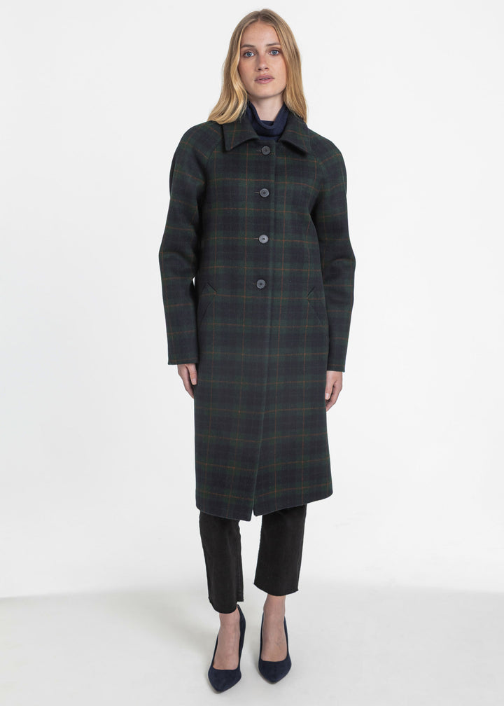 A woman modeling a long dark green plaid coat with black trousers and navy heels, standing against a plain white studio background.