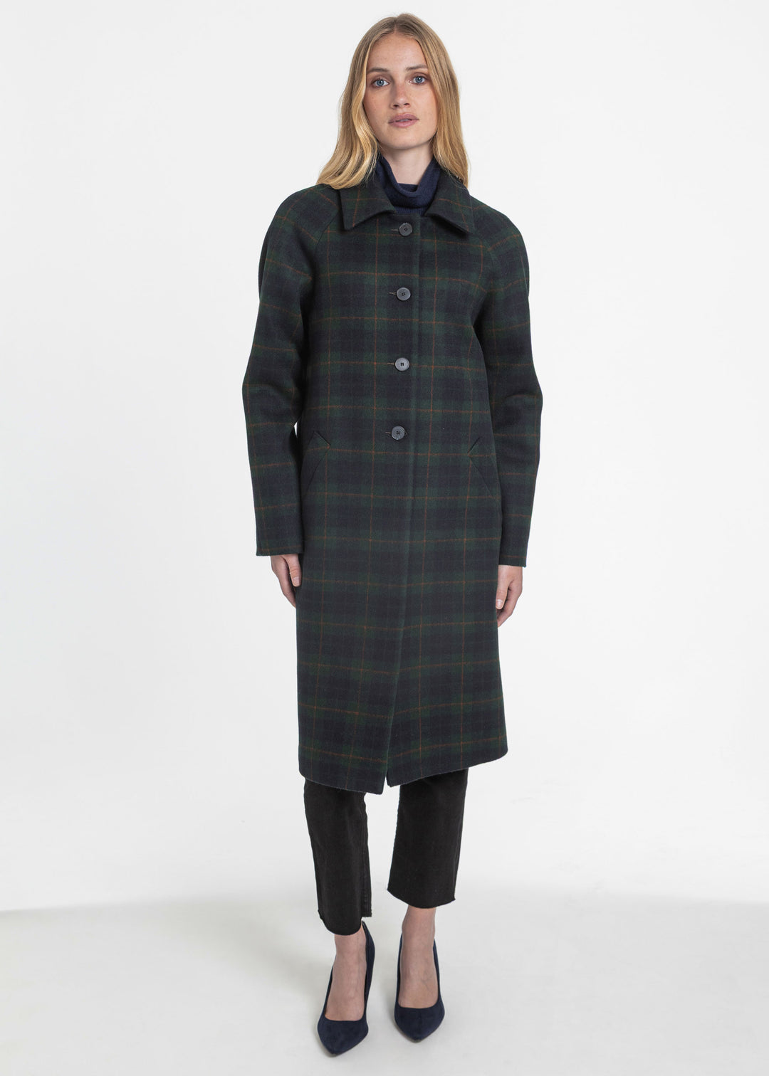 A woman modeling a long dark green plaid coat with black trousers and navy heels, standing against a plain white studio background.