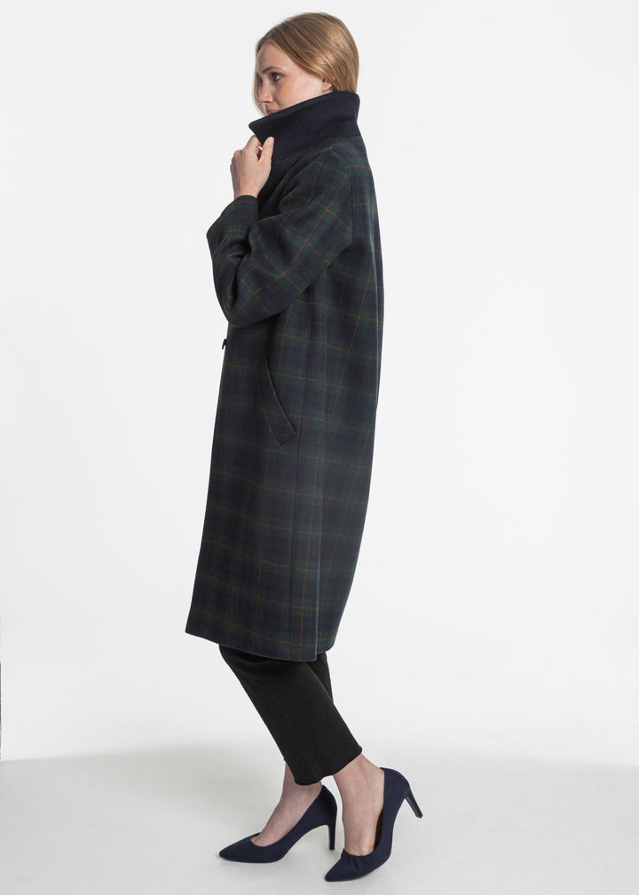 A woman modeling a long dark green and navy plaid coat with a high dark collar, paired with black pants and navy heels, against a plain white studio background.